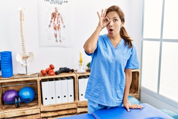 Middle age hispanic physiotherapist woman working at pain recovery clinic doing ok gesture shocked...