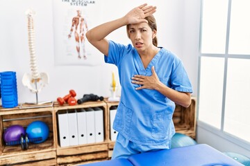 Middle age hispanic physiotherapist woman working at pain recovery clinic touching forehead for illness and fever, flu and cold, virus sick