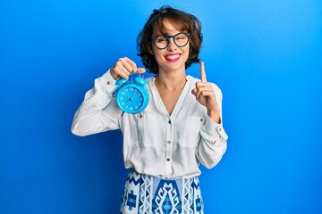 Young brunette woman holding alarm clock smiling with an idea or question pointing finger with happy face, number one