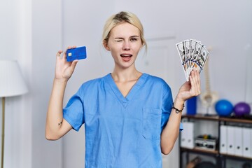 Young physiotherapist woman holding credit card and dollars banknotes winking looking at the camera...