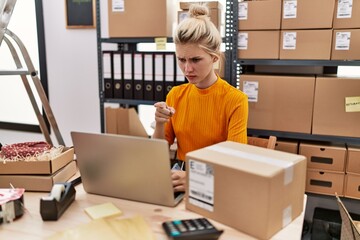 Young blonde woman working at small business ecommerce using laptop pointing with finger to the camera and to you, confident gesture looking serious