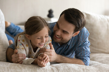 Happy young caring father cuddling laughing little child daughter, having fun or enjoying pleasant funny trustful conversation, lying together on comfortable sofa, loving family relationship concept.