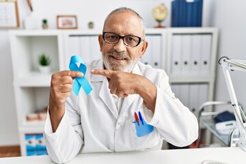 Mature doctor man holding blue ribbon at the clinic smiling happy pointing with hand and finger