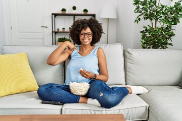 Obraz premium African american woman with afro hair sitting on the sofa eating popcorn at home pointing finger to one self smiling happy and proud