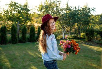 a red-haired teenage girl in a red hat is dancing on the lawn with a bouquet of flowers. the setting sun