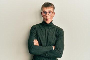 Young caucasian man wearing glasses with arms crossed gesture making fish face with mouth and squinting eyes, crazy and comical.