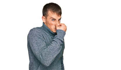 Young blond man wearing casual clothes smelling something stinky and disgusting, intolerable smell, holding breath with fingers on nose. bad smell