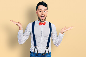 Hispanic man with beard wearing hipster look with bow tie and suspenders celebrating crazy and...