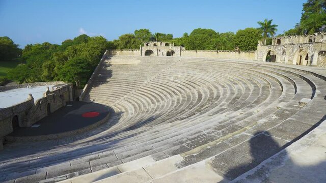 Semicircular rows of seats rise above the stage. The old walls of the amphitheater are flooded with the morning sun. Tropical vegetation, ancient walls and blue skies create a beautiful landscape.