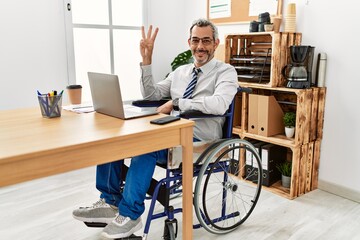 Middle age hispanic man working at the office sitting on wheelchair showing and pointing up with fingers number three while smiling confident and happy.