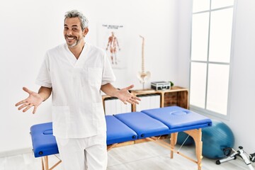 Middle age hispanic therapist man working at pain recovery clinic smiling cheerful with open arms as friendly welcome, positive and confident greetings