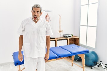 Obraz premium Middle age hispanic therapist man working at pain recovery clinic in shock face, looking skeptical and sarcastic, surprised with open mouth