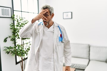 Fototapeta premium Middle age hispanic man wearing doctor uniform and stethoscope at waiting room peeking in shock covering face and eyes with hand, looking through fingers with embarrassed expression.