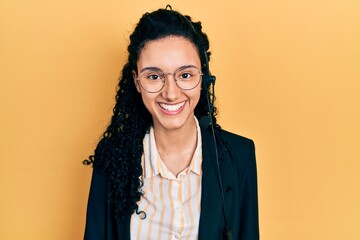 Young hispanic woman with curly hair wearing call center agent headset looking positive and happy standing and smiling with a confident smile showing teeth