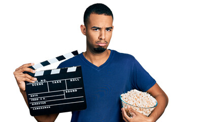 Young african american man eating popcorn holding film clapboard depressed and worry for distress,...