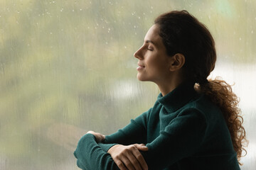 Music of rain. Side shot of serene latin woman relax by window with closed eyes listen sound of raindrops feel pleasure. Calm young lady enjoy breathing fresh ozonized air after rainstorm. Copy space. - Powered by Adobe