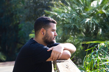 A gay man of Latin origin, dark and bearded, stands pensively looking nowhere in the park. He is...
