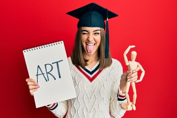 Young brunette girl graduated on art sticking tongue out happy with funny expression.