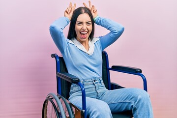 Beautiful woman with blue eyes sitting on wheelchair posing funny and crazy with fingers on head as...