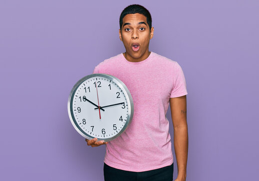 Young handsome hispanic man holding big clock scared and amazed with open mouth for surprise, disbelief face