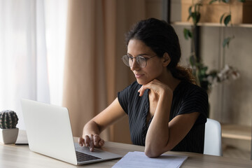 Interesting task. Focused young hispanic woman in eyewear sit by desk at home work online by laptop...