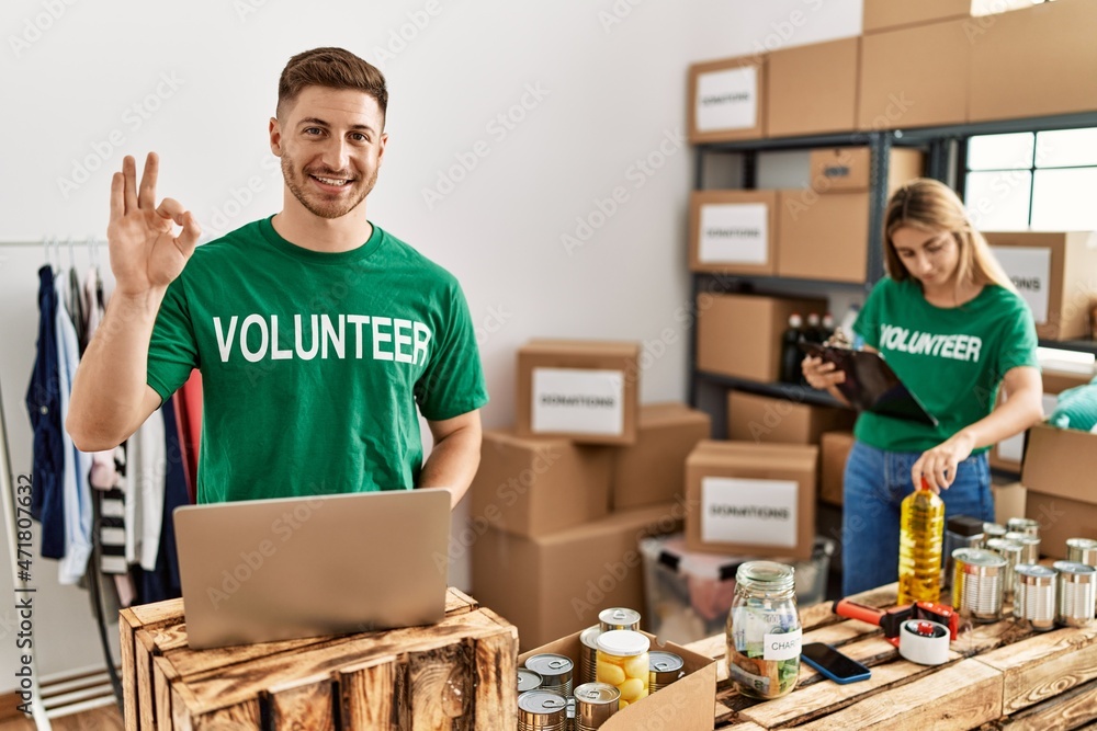 Sticker young man and woman wearing volunteer t shirt at donations stand doing ok sign with fingers, smiling