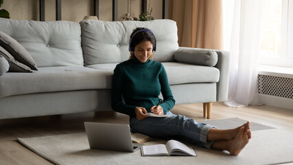 Interested in learning. Latina female student sit on floor surrounded by books use pc listen to...
