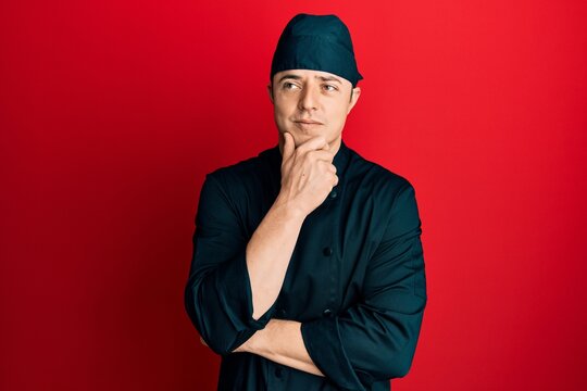 Handsome young man wearing professional cook uniform and hat with hand on chin thinking about question, pensive expression. smiling with thoughtful face. doubt concept.