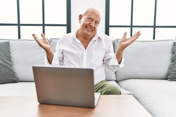 Senior man using laptop at home sitting on the sofa clueless and confused expression with arms and...