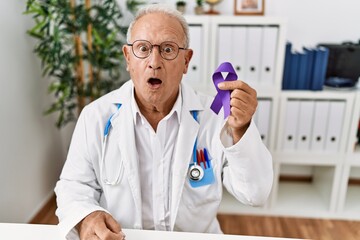 Senior doctor man holding purple ribbon awareness scared and amazed with open mouth for surprise,...