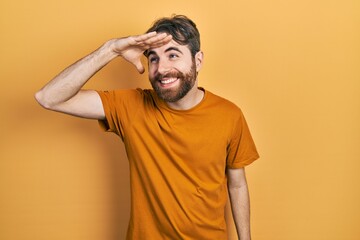 Fototapeta na wymiar Caucasian man with beard wearing casual yellow t shirt very happy and smiling looking far away with hand over head. searching concept.
