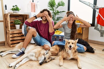Young hispanic couple doing laundry with dogs covering eyes with hands smiling cheerful and funny....