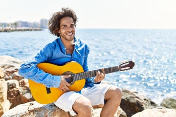 Young hispanic man playing classical guitar sitting on rock at the beach.