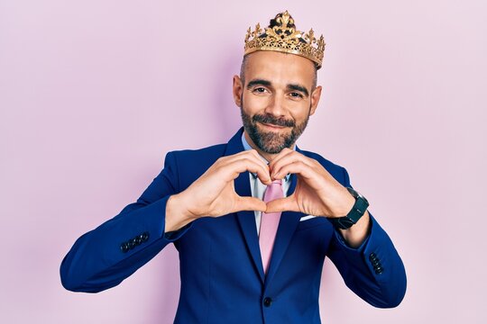 Young hispanic man wearing business clothes and king crown smiling in love doing heart symbol shape with hands. romantic concept.