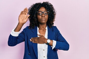 Young african american woman wearing business clothes and glasses swearing with hand on chest and...
