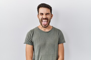 Young hispanic man with beard wearing casual t shirt over white background sticking tongue out happy with funny expression. emotion concept.
