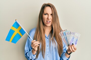 Young blonde woman holding sweden flag and krone banknotes in shock face, looking skeptical and sarcastic, surprised with open mouth