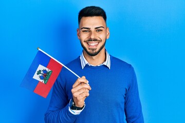 Young hispanic man with beard holding haiti flag looking positive and happy standing and smiling with a confident smile showing teeth