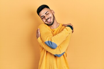 Young hispanic man with beard wearing casual winter sweater hugging oneself happy and positive,...