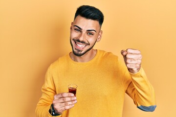 Young hispanic man with beard drinking whiskey shot screaming proud, celebrating victory and success very excited with raised arms