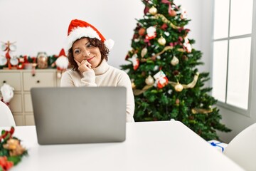 Middle age hispanic woman using laptop sitting by christmas tree serious face thinking about question with hand on chin, thoughtful about confusing idea