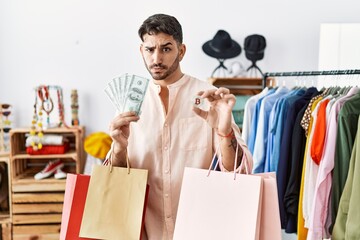 Young hispanic man holding shopping bags, dollars and bitcoin skeptic and nervous, frowning upset...