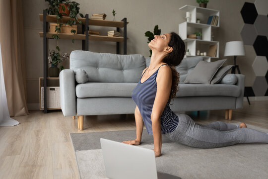 Strengthening muscles. Fit young hispanic woman do yoga exercises on floor before computer screen. Attractive millennial female practice cobra pose stretch shoulders and back reduce stress breath deep