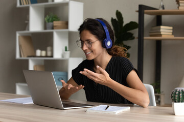 Fototapeta na wymiar Confident hispanic female interpreter work from home office sit at desk in headset talk look on laptop screen translate online event. Young lady participate at web conference negotiate using pc webcam