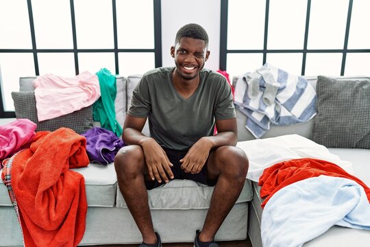 Young african american man sitting on the sofa with dirty laundry clothes winking looking at the camera with sexy expression, cheerful and happy face.