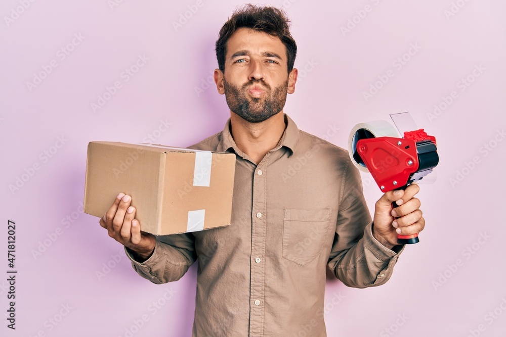 Sticker Handsome man with beard holding packing tape holding cardboard looking at the camera blowing a kiss being lovely and sexy. love expression.