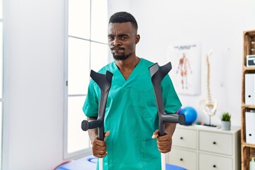 Young african physiotherapist man holding crutches skeptic and nervous, frowning upset because of...