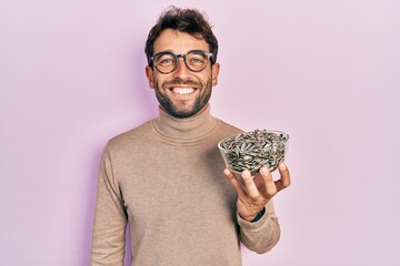 Handsome man with beard football reporter holding pumpkin seeds bowl looking positive and happy standing and smiling with a confident smile showing teeth - obrazy, fototapety, plakaty