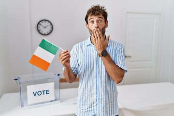 Young handsome man at political campaign election holding ireland flag covering mouth with hand,...
