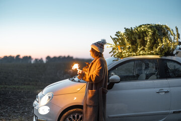 Happy caucasian woman firing sparklers near car with Christmas tree on nature at dusk. Concept of celebrating New Year holidays. Idea of Christmas mood and fun. Side view with sunset sky on background